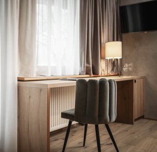 Peternhof_Suite_Schreibtisch_Fensterfront.jpg Holzschreibtisch mit gepolstertem Samtstuhl vor einer Fensterfront mit fließenden Vorhängen, daneben eine Tischleuchte mit warmem Licht.